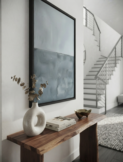 Affordable minimalist abstract wall decor with premium textured canvas by Noho Art Gallery with a black frame hangs on a white wall above a rustic wooden console table. The artwork features a textured, painterly design in shades of gray and blue.