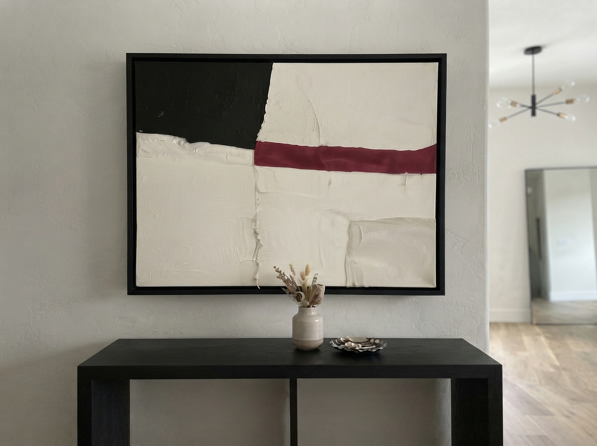 A framed Minimalist black and white abstract canvas with red accent, minimalist abstract painting with red accent by Noho Art Gallery with black, off-white, and burgundy geometric blocks hangs above a dark console table in a modern interior setting.
