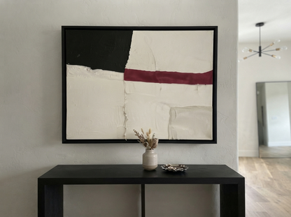 A framed Minimalist black and white abstract canvas with red accent, minimalist abstract painting with red accent by Noho Art Gallery with black, off-white, and burgundy geometric blocks hangs above a dark console table in a modern interior setting.