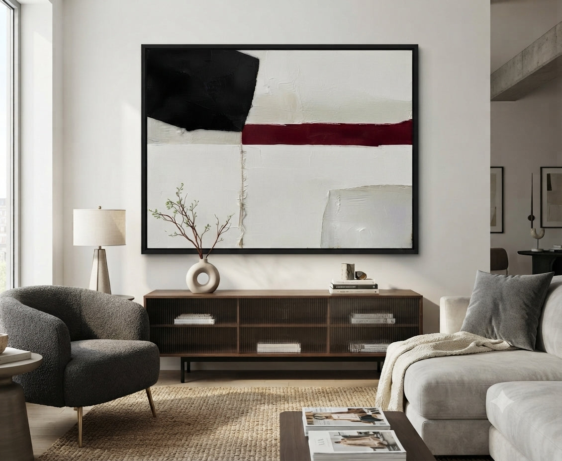 A large Minimalist black and white abstract canvas with red accent, large black and white textured painting with red highlight by Noho Art Gallery hangs above a dark wood console in a modern living room. The artwork features a white background with a bold black block, a horizontal dark red stripe, and soft gray brushstrokes.