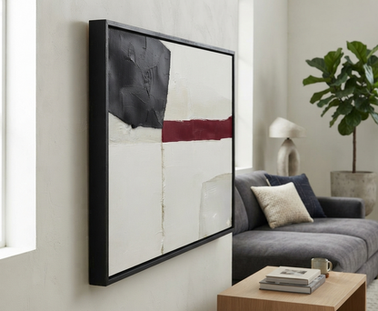 A framed Minimalist black and white abstract canvas with red accent, bold minimalist wall art original acrylic on canvas by Noho Art Gallery hangs on a light beige wall in a modern living room setting, featuring a black frame, a white background, a large black textured shape, and a horizontal dark red stripe.