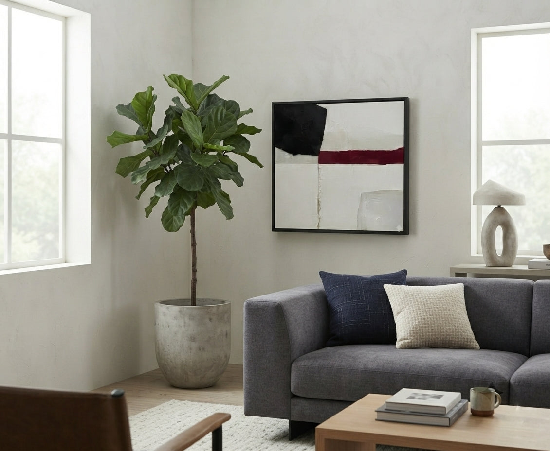 A modern living room scene featuring a large potted fiddle-leaf fig plant in a textured concrete pot, positioned next to a gray fabric sofa with navy and cream pillows. On the wall above the sofa is a Minimalist black and white abstract canvas with red accent, statement piece abstract painting black white and crimson by Noho Art Gallery with black, white, and red geometric blocks.