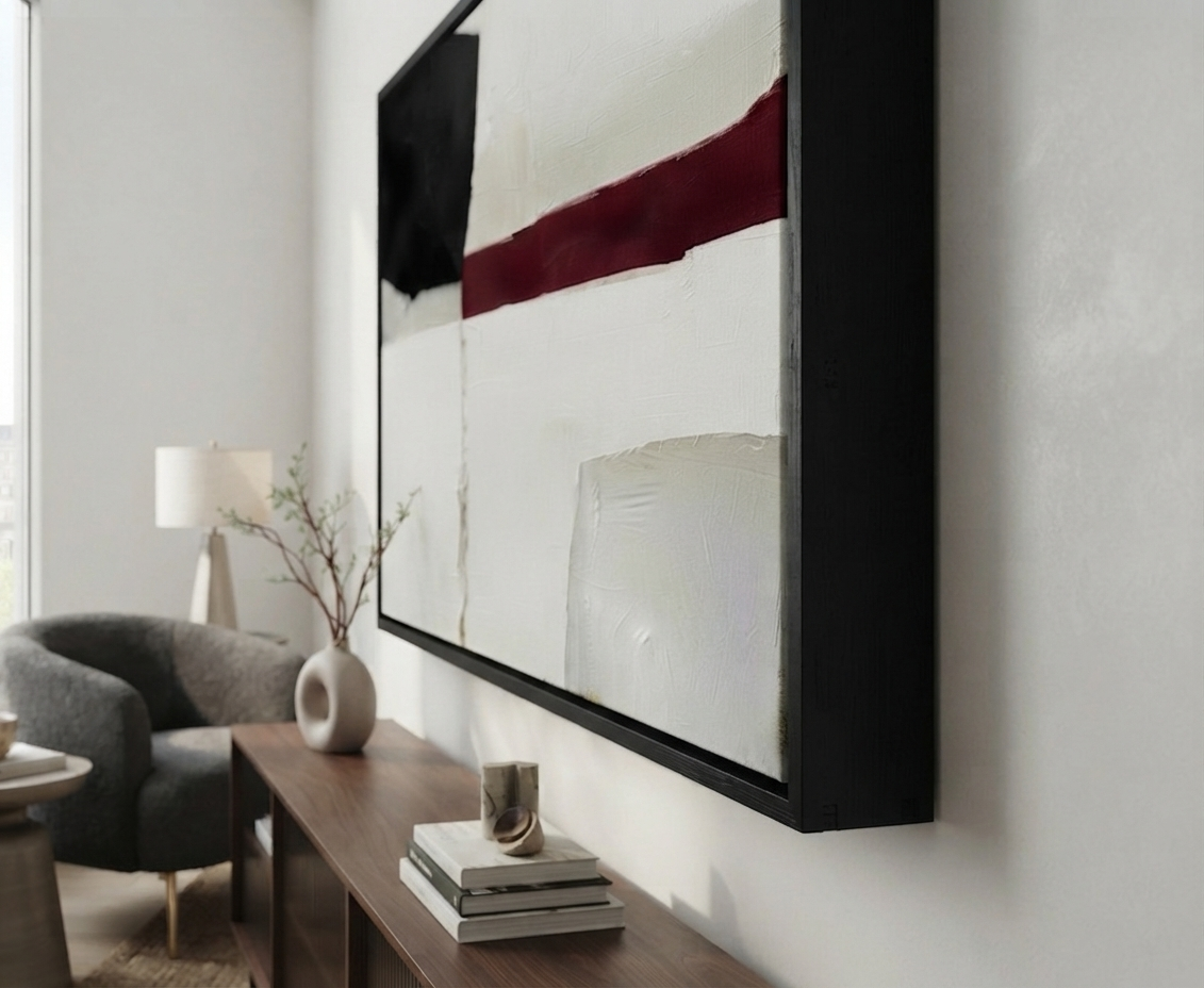 A large Minimalist black and white abstract canvas with red accent, large black and white textured painting with red highlight by Noho Art Gallery hangs on a light gray wall above a wooden console table in a modern living room setting. The artwork features a bold abstract pattern with white, black, and deep burgundy sections, surrounded by a thick black frame.