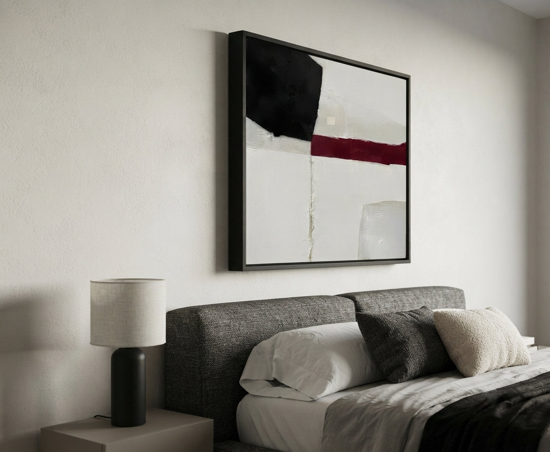 A modern bedroom scene featuring a framed Minimalist black and white abstract canvas with red accent, minimalist geometric abstract painting for modern home decor by Noho Art Gallery with black, white, and red geometric blocks above a bed with gray and white bedding. A bedside table with a black lamp is visible on the left.