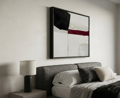 A modern bedroom scene featuring a framed Minimalist black and white abstract canvas with red accent, minimalist geometric abstract painting for modern home decor by Noho Art Gallery with black, white, and red geometric blocks above a bed with gray and white bedding. A bedside table with a black lamp is visible on the left.