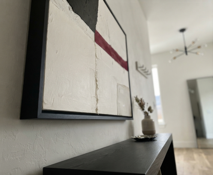 A framed Minimalist black and white abstract canvas with red accent, contemporary black and white art with pop of color by Noho Art Gallery with textured off-white, black, and red brushstrokes hanging on a white wall above a dark console table in a modern interior.