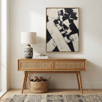 A mid-century modern wooden console table with woven cane panels, styled in a bright entryway with a lamp, books, a basket, and a Custom black and white statement piece painting for luxury interiors by Noho Art Gallery on the wall.