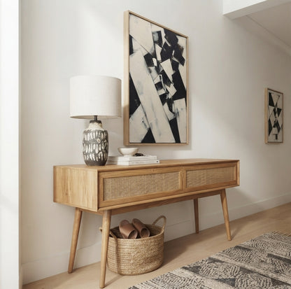 A mid-century modern wooden console table with woven cane drawer fronts, styled in a bright room with a patterned Minimalist black and white acrylic painting on canvas with modern geometric details by Noho Art Gallery, a ceramic table lamp, books, and a woven basket underneath.