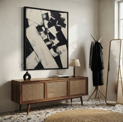 A large framed Original monochrome geometric canvas art with bold architectural composition by Noho Art Gallery piece hangs above a mid-century modern wooden sideboard in a styled living room with a mirror, coat rack, lamp, and textured rug.