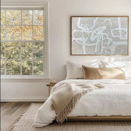 Neatly made bed with beige and white bedding in a room with a large window and abstract wall art.