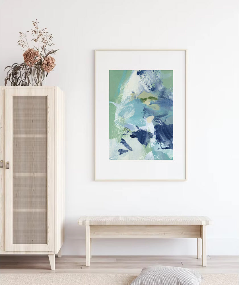 Framed abstract art piece on a white wall above a wooden bench, with a cabinet and plant to the left.