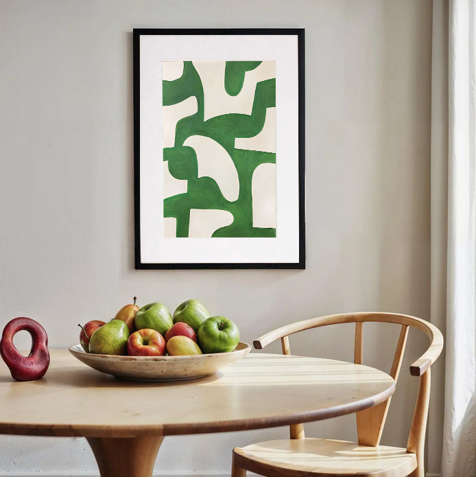 Framed abstract art with green and beige patterns on a wall above a table with apples.